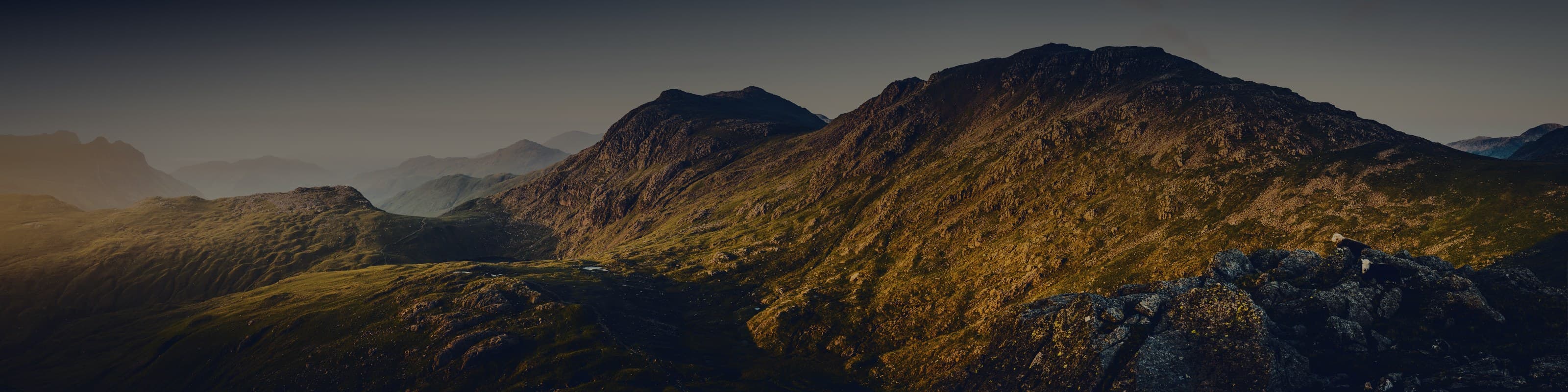 The Lakeland fells at sunrise, seen from a neighbouring top: a dark valley on the left, and on the right a long ridge climbing to a summit, its crest lit gold by the low sun.